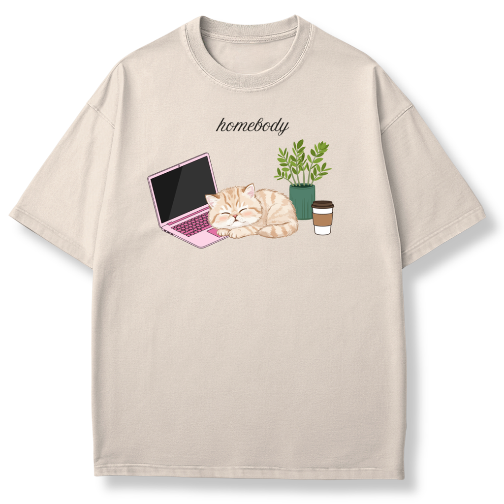 A light beige crewneck t-shirt featuring a cute graphic of a sleeping orange tabby cat next to a pink laptop, a green potted plant, and a takeaway coffee cup, with the word 'homebody' printed above.