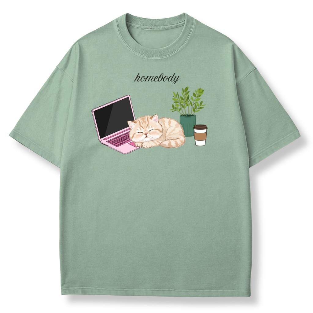 A sage green short-sleeve t-shirt with a cute graphic of a sleeping orange tabby cat next to a pink laptop, a green potted plant, and a coffee cup, with the word 'homebody' printed above.