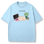 A light blue short-sleeve t-shirt with a graphic print of a sleeping orange tabby cat next to a pink laptop, a potted plant, and a takeaway coffee cup, with the word 'homebody' printed above.